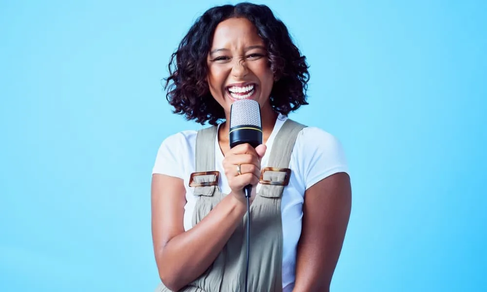 How to Find Your Voice Type and Range 6 Woman with brown curly hair standing in front of a blue background with a microphone practicing her signing ranges and pitches.